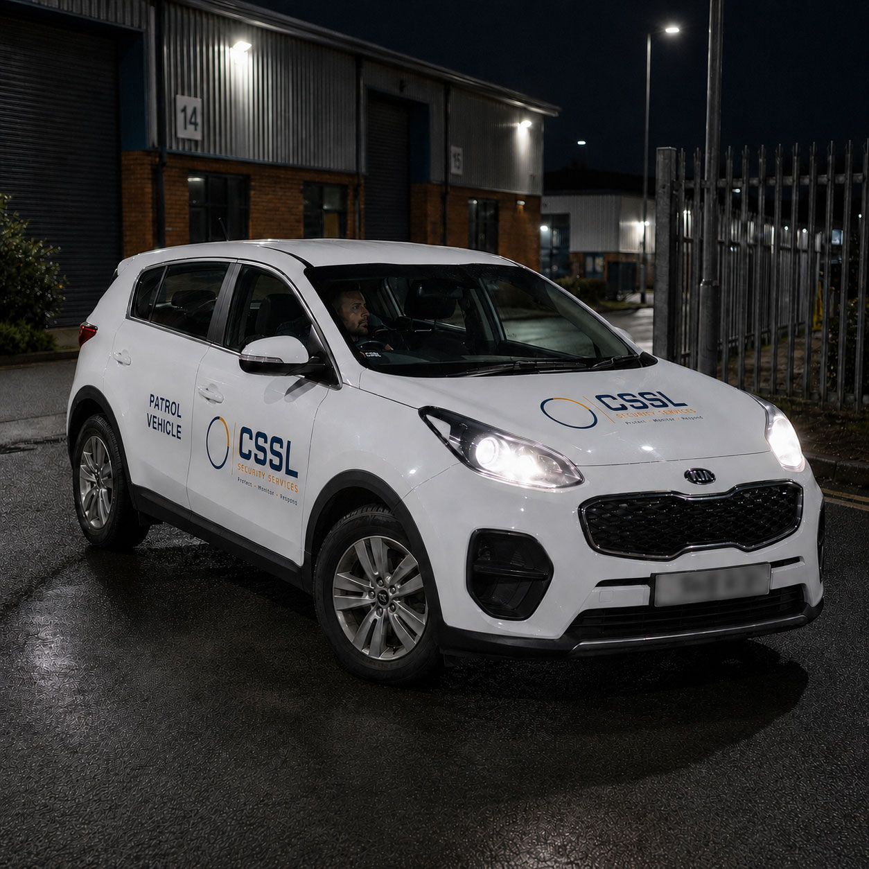 A white CSSL Security Services patrol car with headlights on, parked outside an industrial warehouse building at night with a security guard visible in the driver's seat.